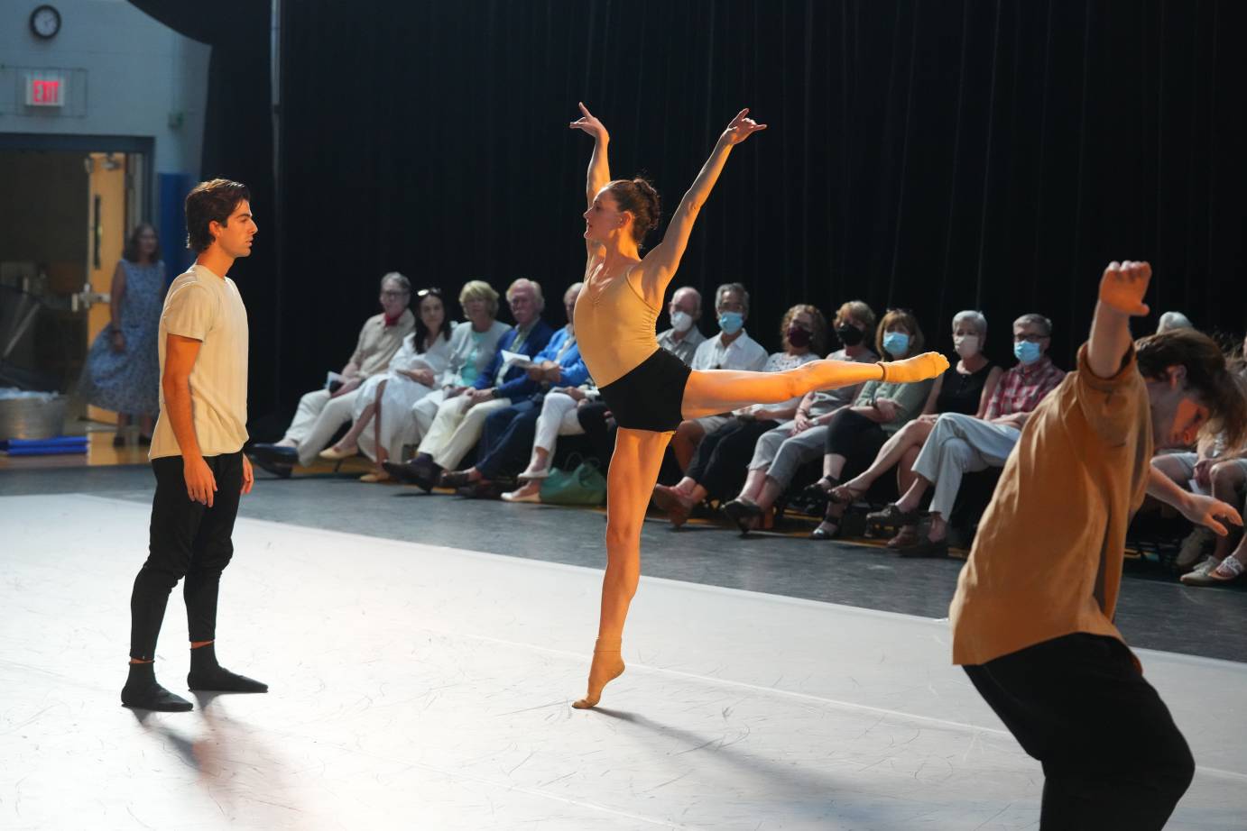 as an audience watches Megan LeCrone a light skinned dancer in a tight gold colored leaotard with fitted black briefs and gold socks darts towards her standing male partner in an arabesque